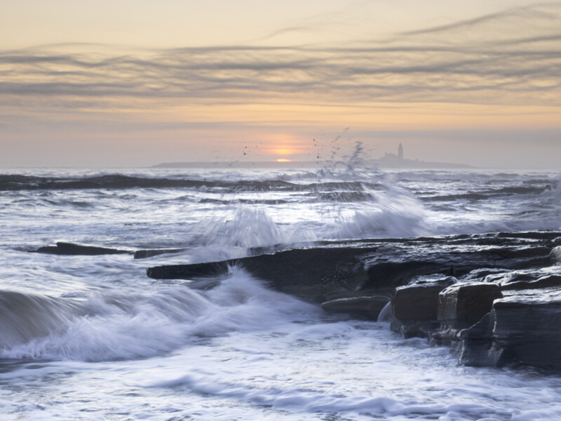 Waves crash against dark rocky outcrops during sunrise, with soft golden light illuminating the misty horizon.