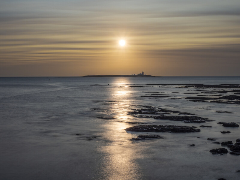 Sun setting over calm ocean waters with rocky foreground and distant landmass silhouetted on horizon.