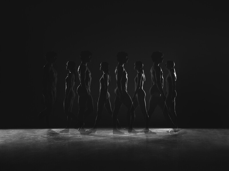 Group of silhouetted figures standing on stage against dark background with dramatic side lighting.