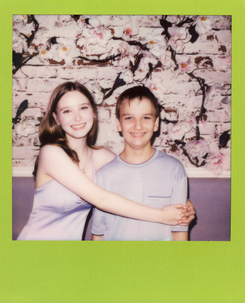 Two young people posing together against a textured floral backdrop with a lime green border.
