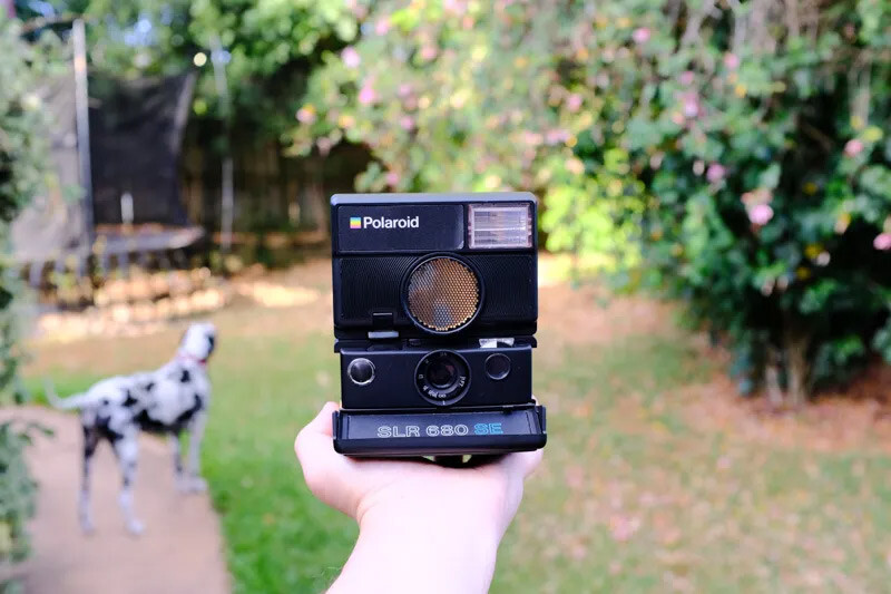 Hand holding a vintage Polaroid SLR 680 instant camera outdoors with a blurred garden background.