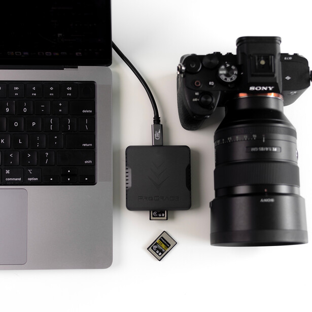 Flat lay of photography and computer equipment including a laptop, Sony camera with lens, external hard drive, SD card, and USB cable on white background.
