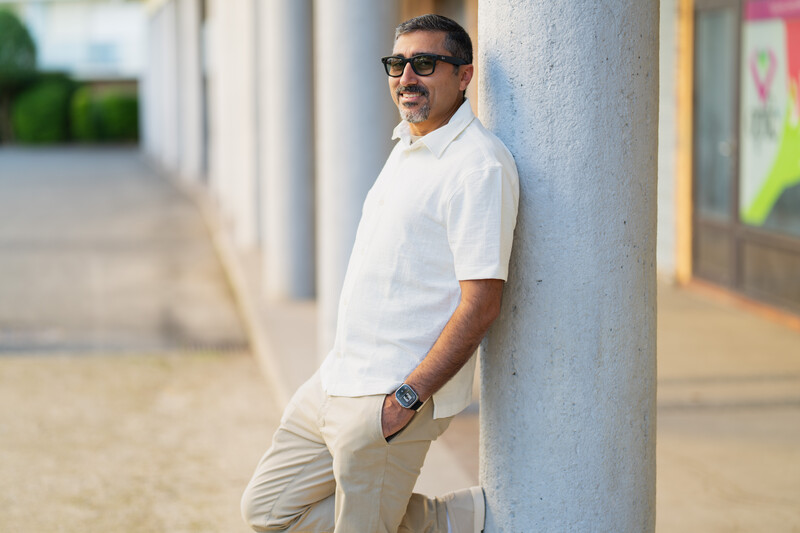 Man in white shirt and neutral pants leaning against a concrete column in a covered corridor.