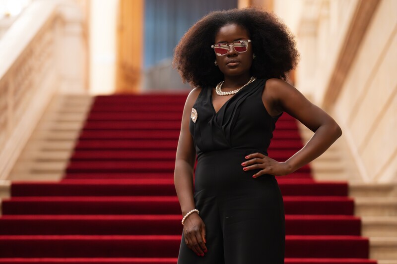 Woman in black sleeveless dress posing on red carpeted staircase in classical architecture setting.
