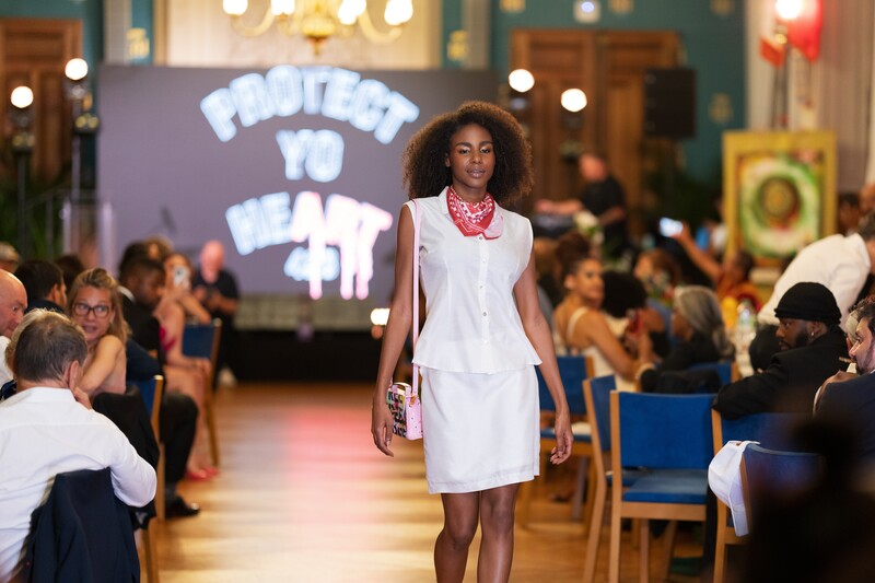 Model wearing white sleeveless dress walks down runway at fashion show with seated audience on both sides.