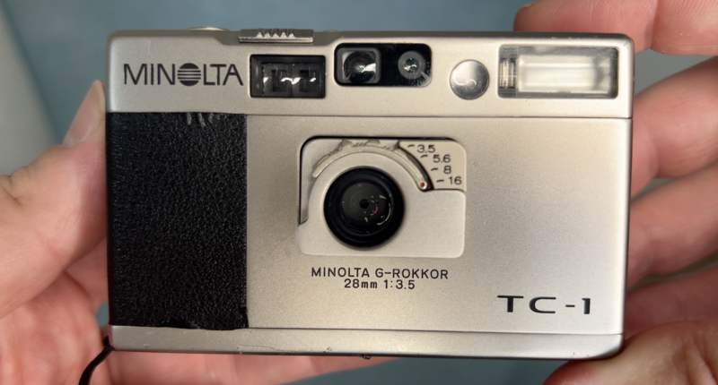Minolta TC-1 compact camera with silver body and black textured grip panel, held in hand.