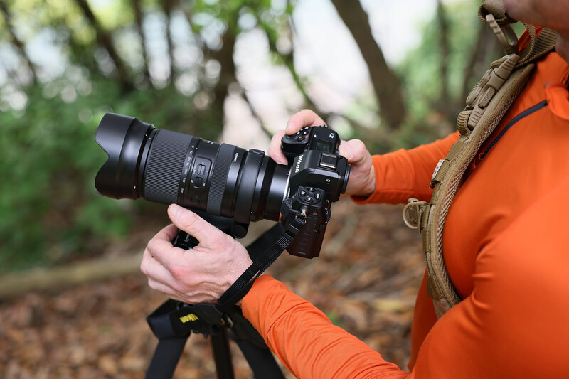 Photographer in orange jacket holding mirrorless camera with telephoto lens on tripod in forest setting.