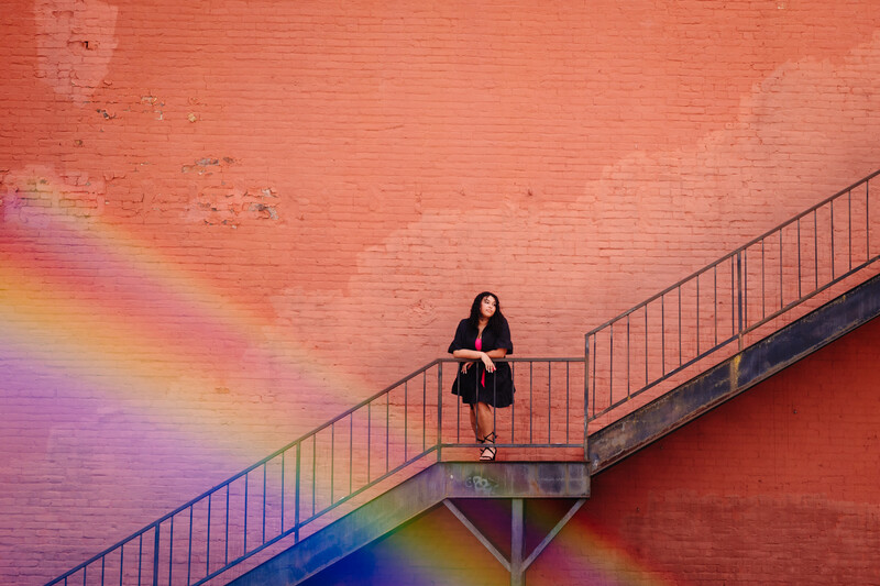 Woman standing on metal platform against vibrant red brick wall with rainbow light projection.