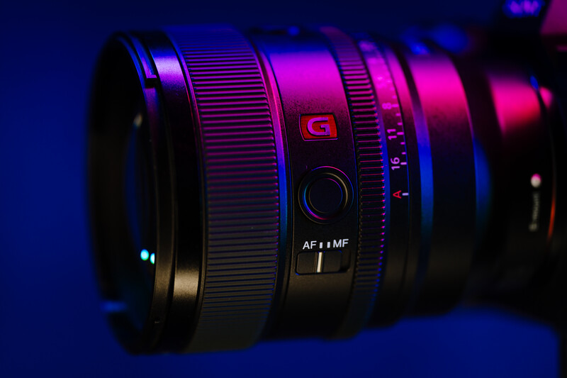 Camera lens with focus ring and autofocus switch illuminated by blue and magenta colored lighting.