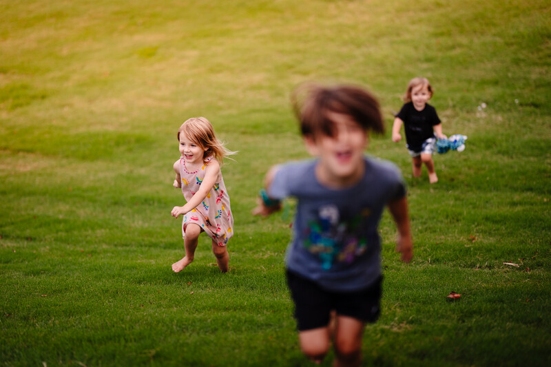 Three young children running across a grassy field, with selective focus on the child in the foreground.