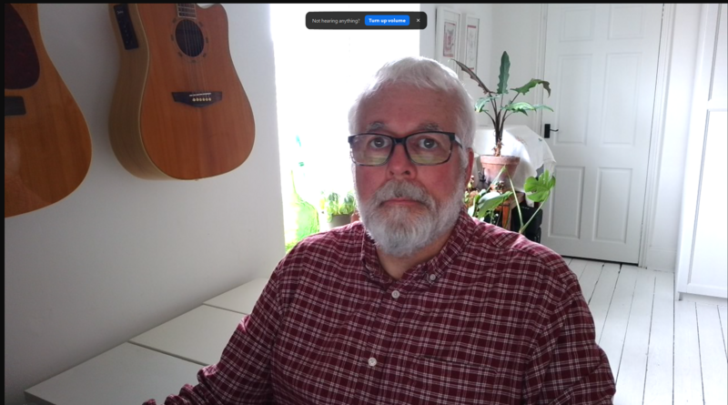 Man with white hair and beard wearing glasses and a burgundy plaid shirt seated indoors.