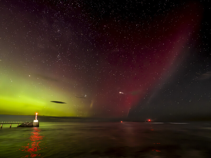 Aurora borealis illuminates a pier and calm water under a starry night sky, casting green and red light across the scene.