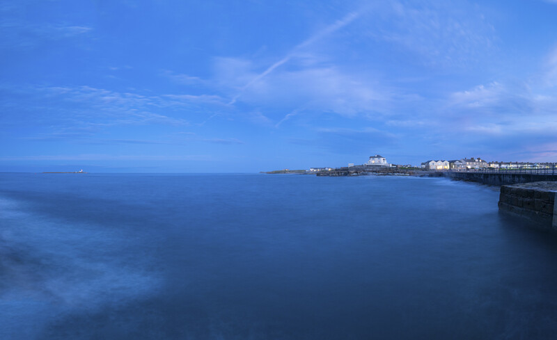 Coastal town at twilight with calm blue water and dark rocky headland in foreground.