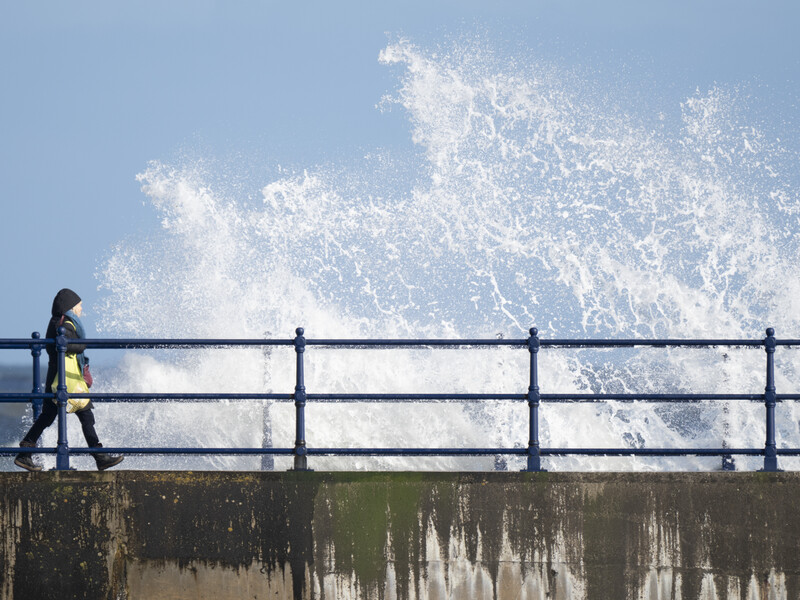 Person in yellow jacket standing on pier railing watching large wave crash against seawall.