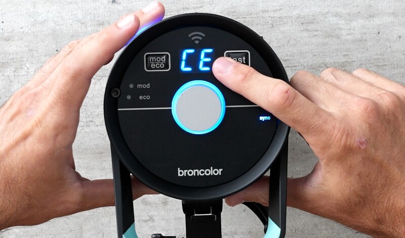Hands holding a round black Broncolor lighting device with blue accents and digital display showing temperature value.