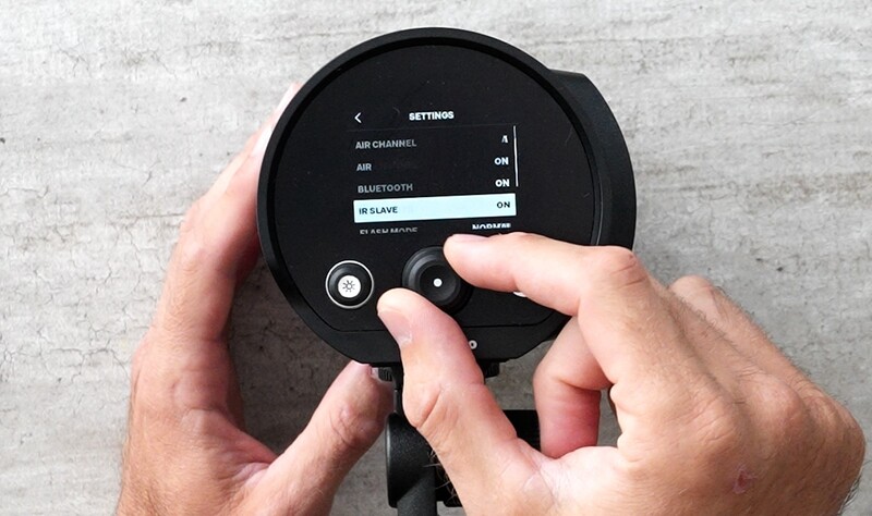 Person holding a round black wireless speaker while navigating the settings menu displayed on its screen.