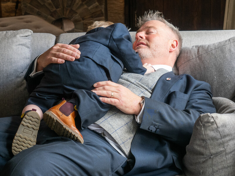 Mature man in navy blazer reclining on gray sofa with eyes closed.