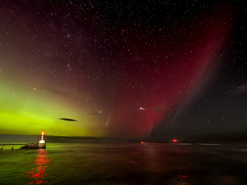 Aurora borealis dancing across night sky above a pier and calm water, illuminated in vibrant green and deep red hues.