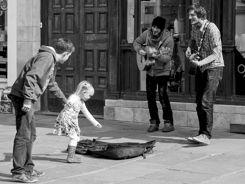 Street musicians performing with guitars while a young child dances on the pavement in front of them.