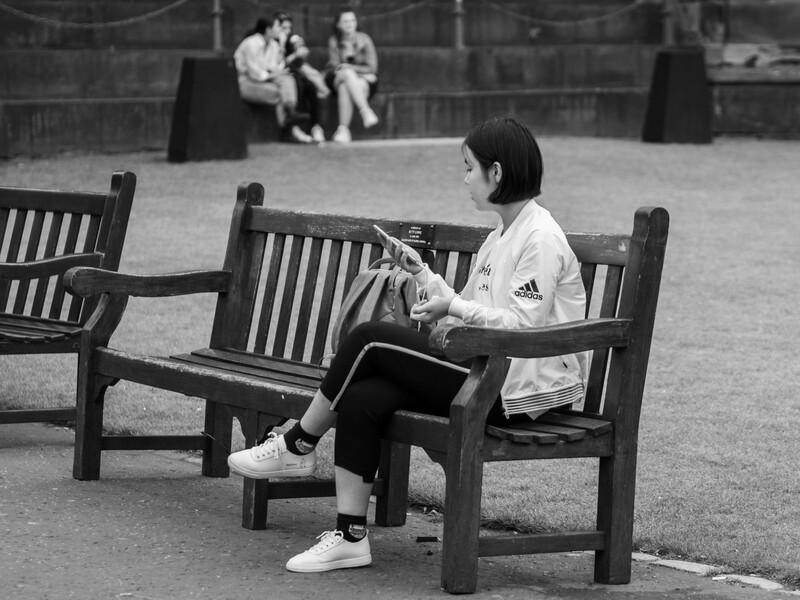 Woman in white Adidas shirt sits reading on a bench, with blurred figures in background on park grounds.