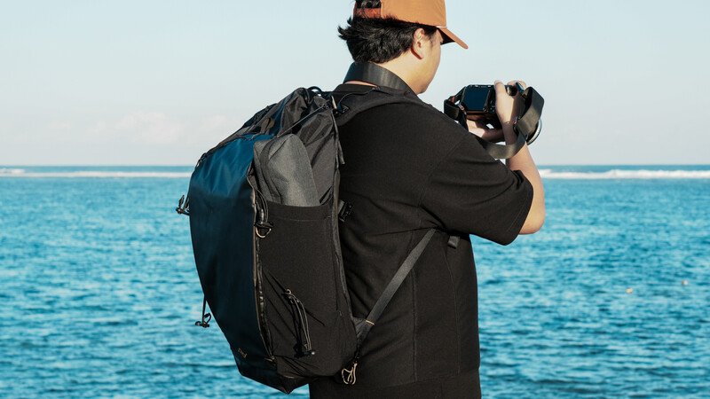 Photographer wearing a large backpack holds a camera to their face while standing on a beach overlooking calm turquoise water.