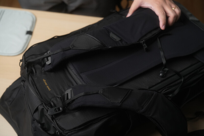 Person holding a black camera backpack with multiple compartments and straps on a wooden table.