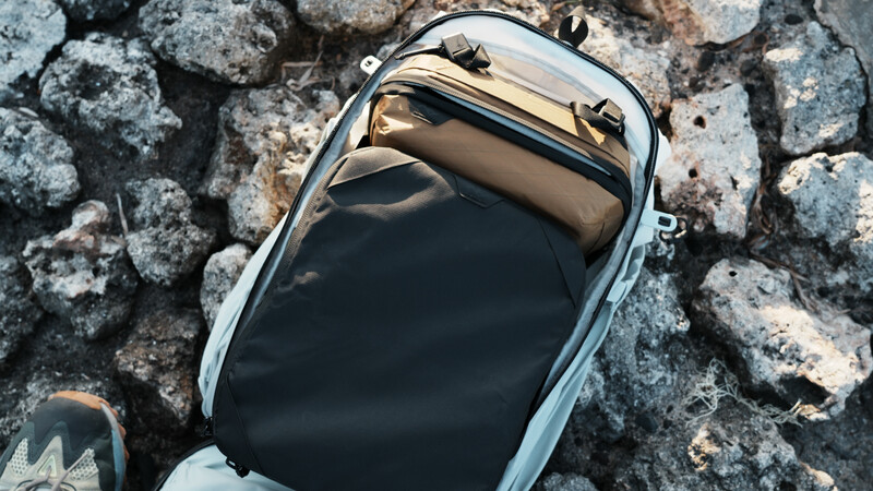 Overhead view of an open camera backpack on rocky ground, revealing tan and black interior compartments.