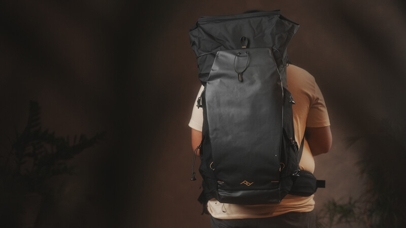 Person wearing a large black roll-top backpack with shoulder straps, photographed from behind against a blurred background.