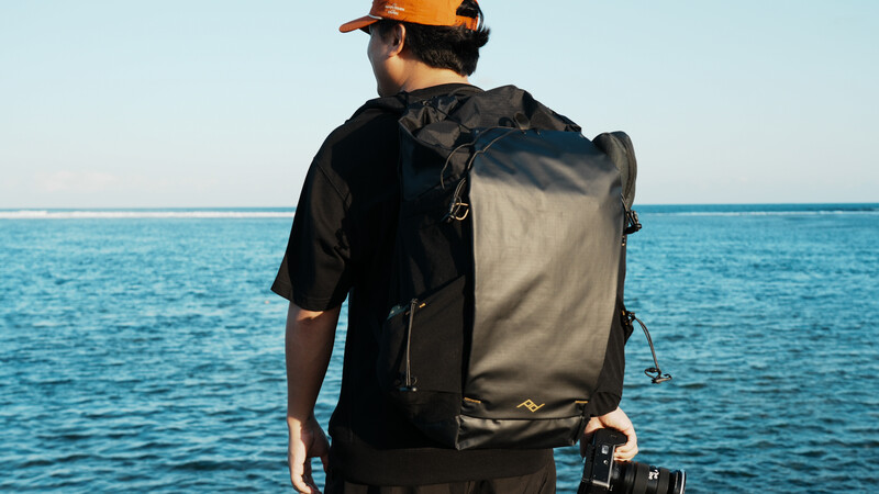 Photographer wearing a waterproof backpack stands facing the ocean with a camera in hand.