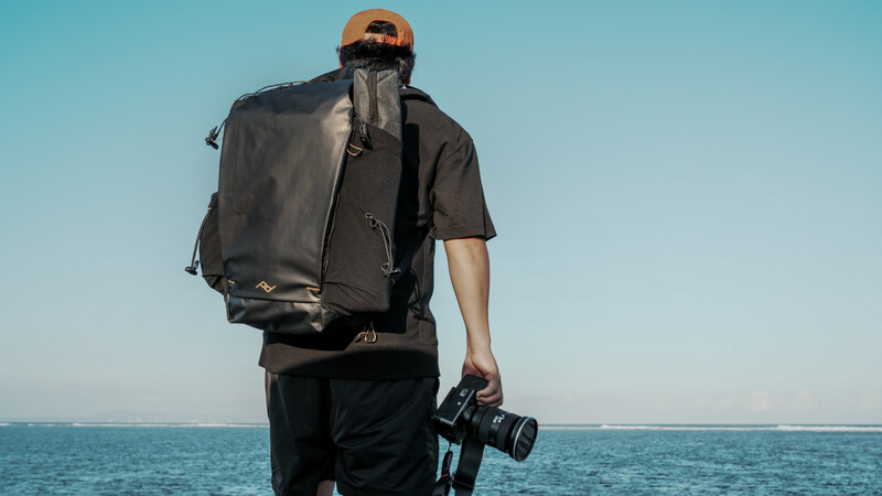 Photographer carrying a large backpack and holding a camera while standing on a beach facing the ocean.