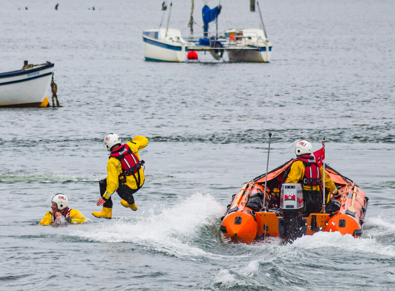 Rescue workers in yellow and red gear guide a person through rough water toward an orange inflatable rescue boat.