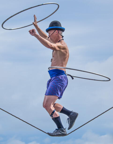 Shirtless male performer jumping while spinning two hula hoops against a blue sky.