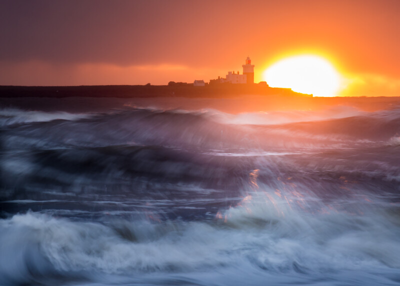 Dramatic coastal sunset with golden sun on horizon, turbulent ocean waves in foreground, silhouetted buildings on distant cliffs.