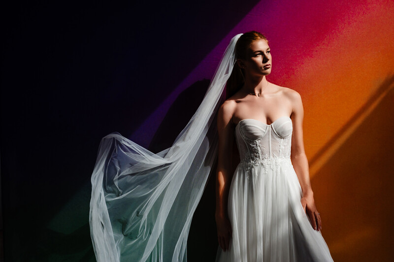 Bride in white strapless gown with flowing veil against vibrant gradient background.