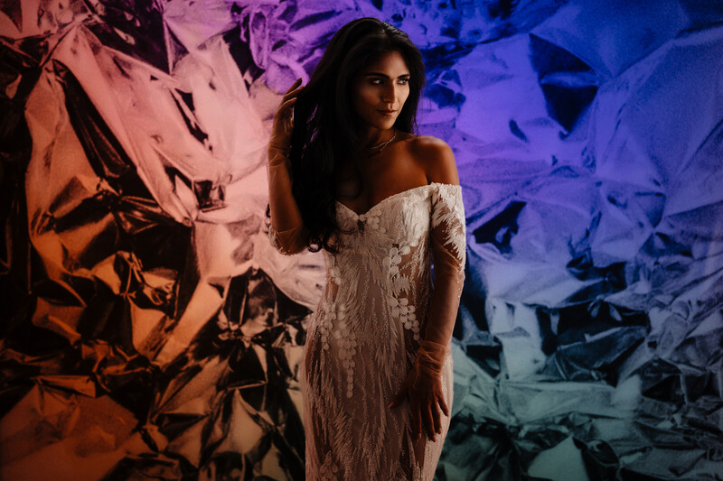 Woman in white off-shoulder dress posed against colorful abstract light backdrop.
