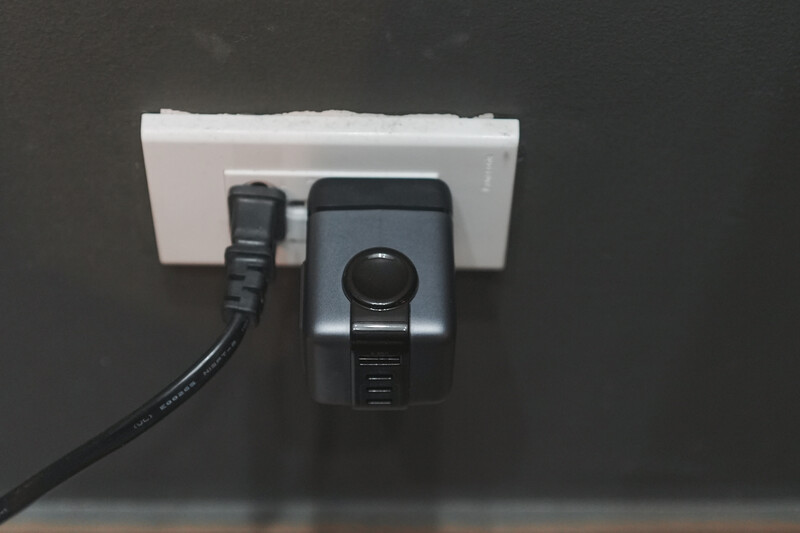 Power outlet mounted on dark wall with black power cord and adapter plugged in.