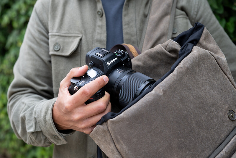 Photographer holding a Nikon mirrorless camera with attached lens, cradled in tan canvas jacket.