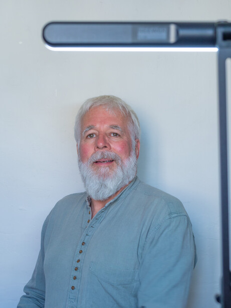 Older man with white beard wearing light blue shirt against white wall with overhead lighting.