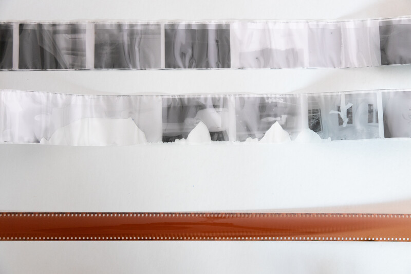 Strips of photographic negatives and film hanging on white string against a white wall above a brown film strip border.