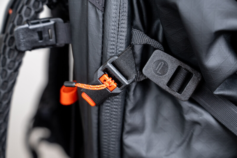 Close-up of black backpack straps with orange plastic clip and gray buckles.