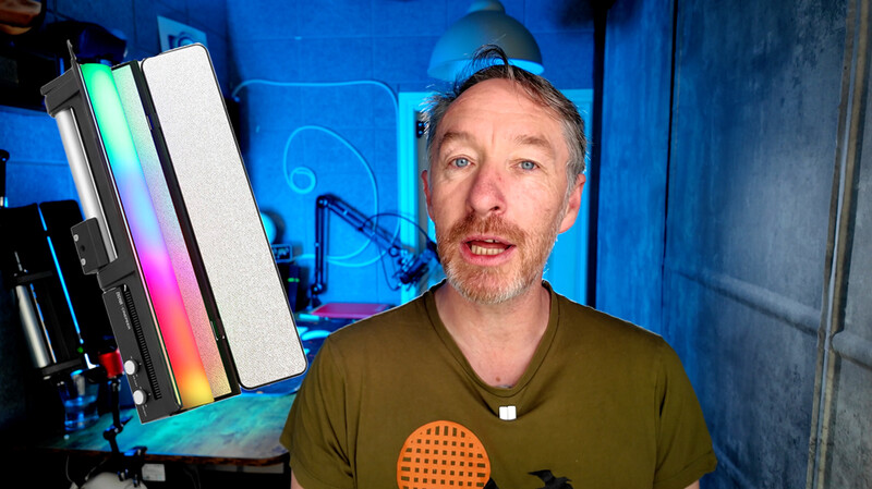 Man in olive shirt speaking to camera in blue-lit studio with RGB lighting panels visible.