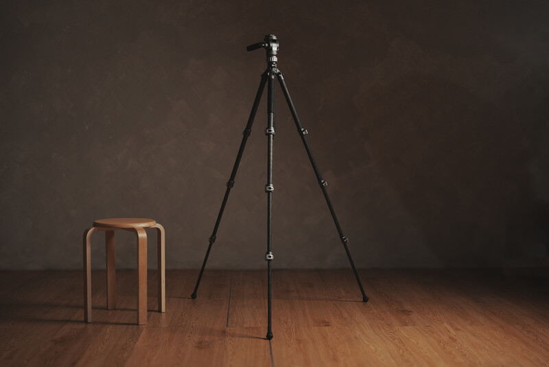 Professional tripod with ball head mounted on wooden floor next to wooden stool against brown wall.