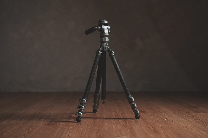 Black tripod with fluid head mounted on wooden floor against neutral background.