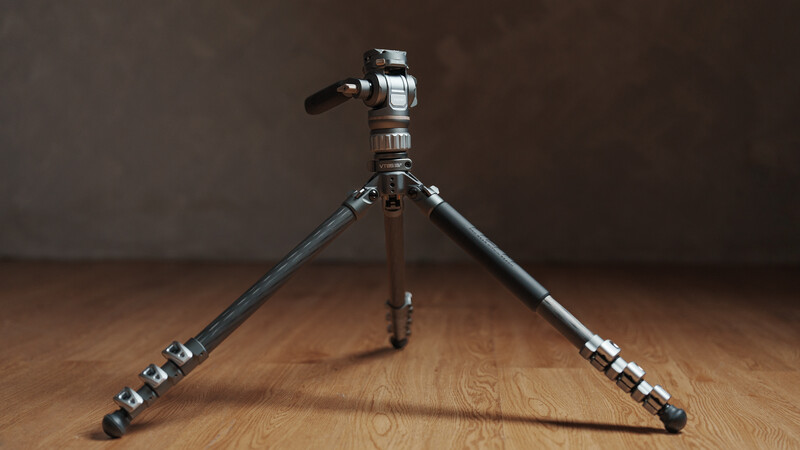 Carbon fiber tripod with ball head mounted on wooden surface against neutral brown backdrop.