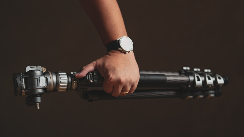 Photographer's hand holding a tripod head with ball mount and quick-release plate, wearing a minimalist watch.