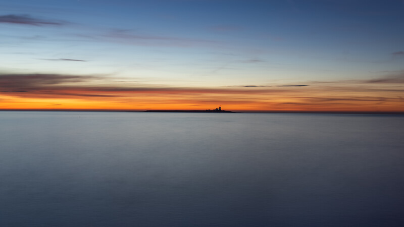Calm ocean horizon at sunset with warm orange and blue gradient sky and distant structure on horizon.