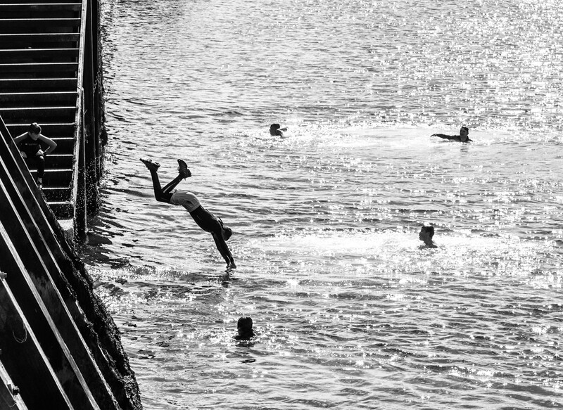 Black and white photograph of multiple people in water near a dock, one figure mid-dive or falling into the water.