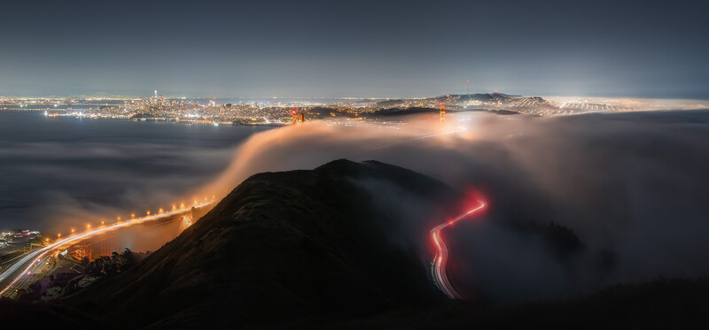 Golden Gate Bridge towers illuminated through thick fog at night, with San Francisco skyline glowing in the background.