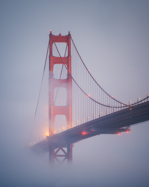 Red suspension bridge shrouded in fog at dawn, with warm lighting visible on the lower tower.