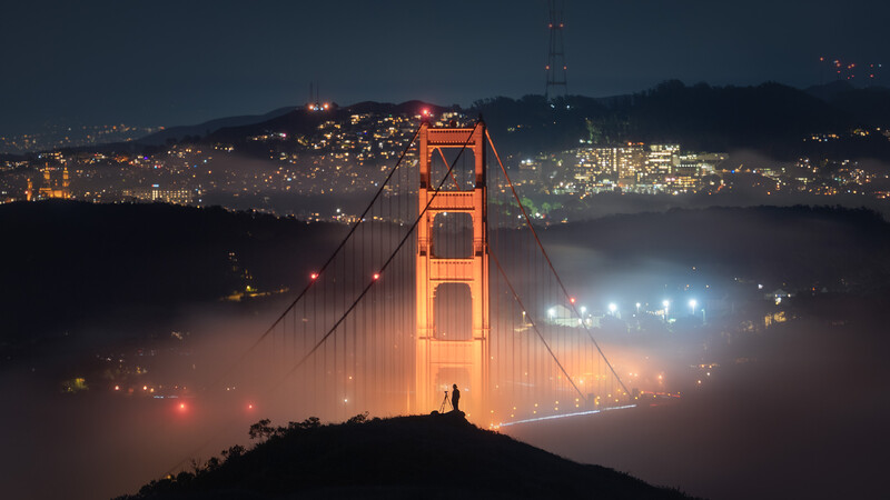 Golden suspension bridge tower rising through fog at night with city lights and silhouetted hills in background.